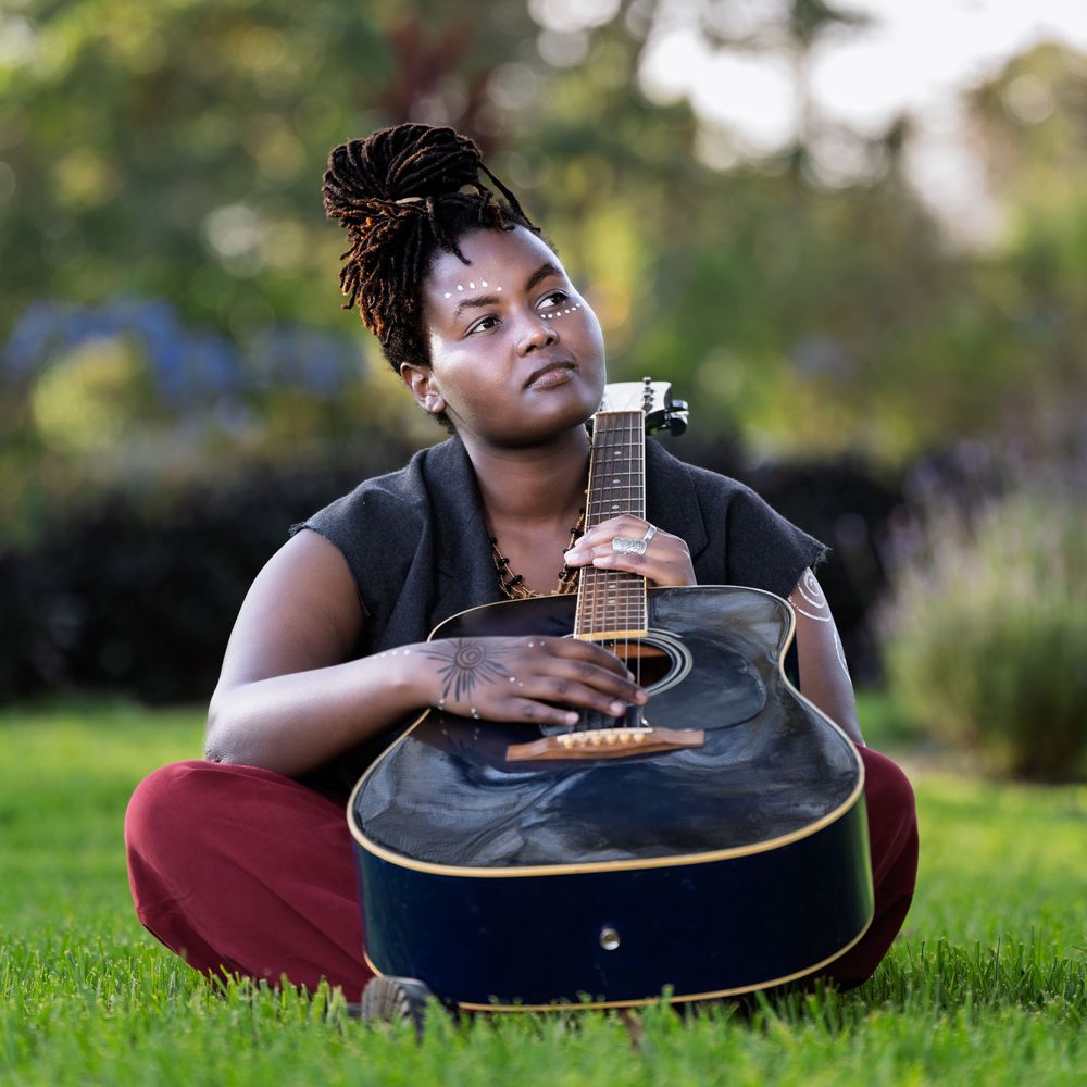 A woman with locs sits cross-legged on grass cradling a blue acoustic guitar in a serene outdoor setting.