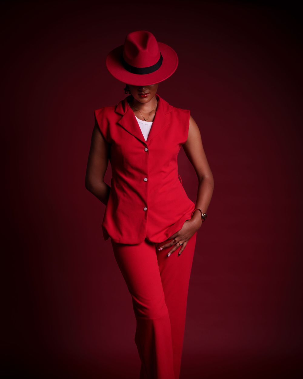 Fashionable woman in red suit and wide-brim fedora poses confidently on dark red background.