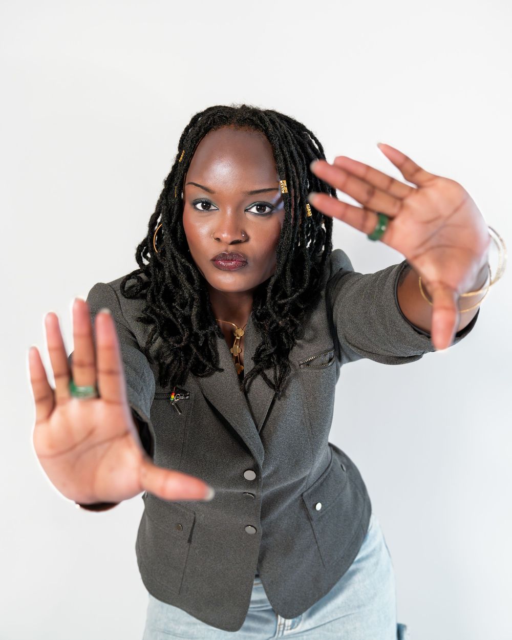 Black woman with locs extending both hands toward camera wearing gray blazer and light jeans on white background.