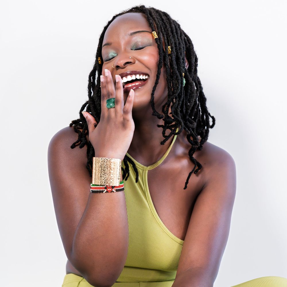 Joyful Black woman with locs laughing with hand to face, wearing gold jewelry and olive green top on white background.