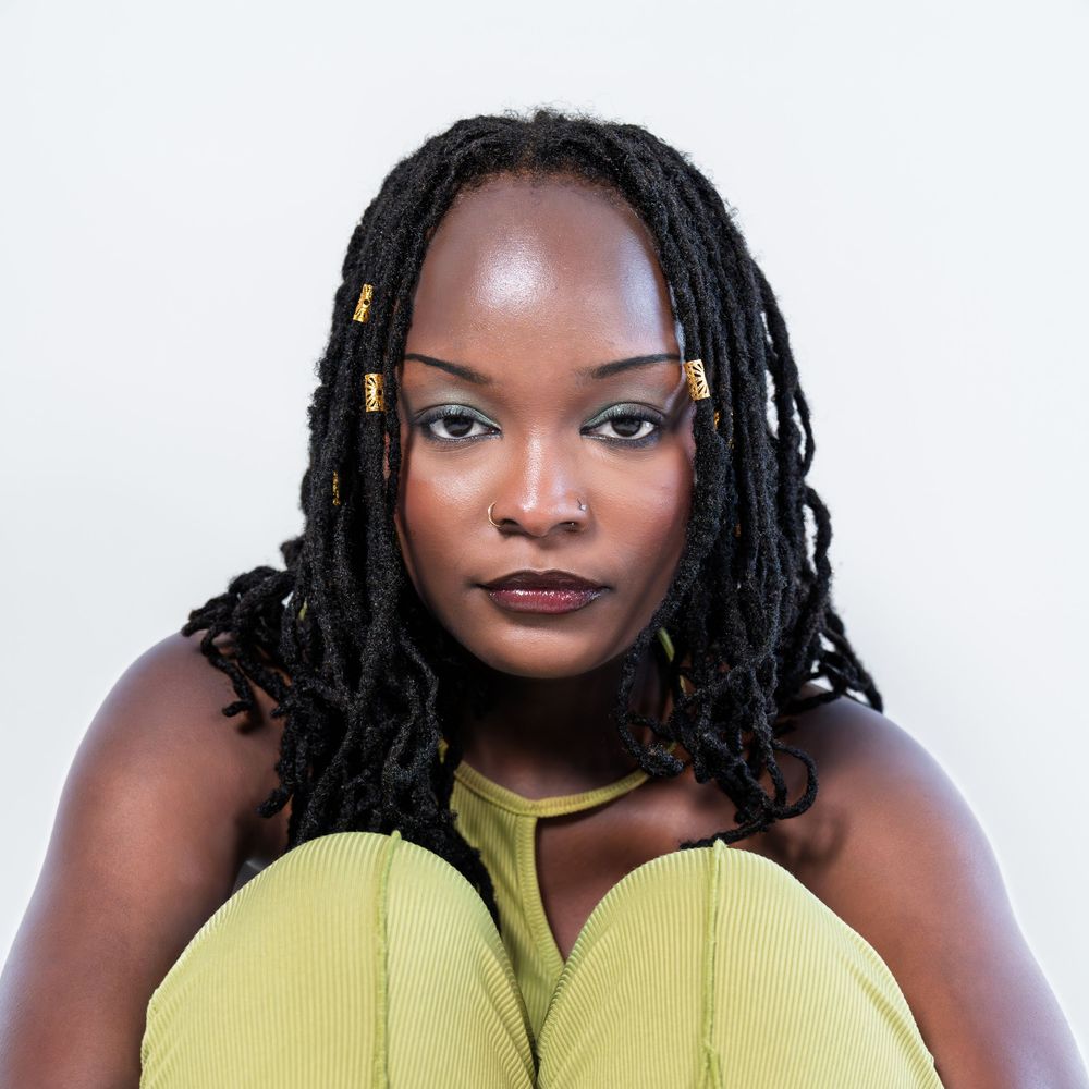 A woman with beaded black locs sits cross-legged in a yellow outfit, posing against a white background.