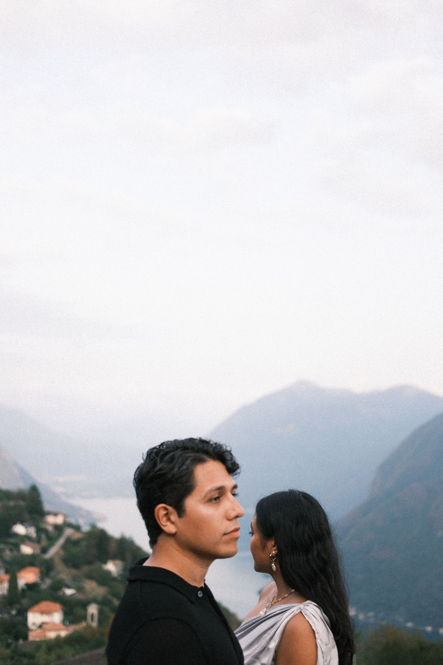 A young man and woman stand close together, gazing in opposite directions, in a soft, muted outdoor setting.