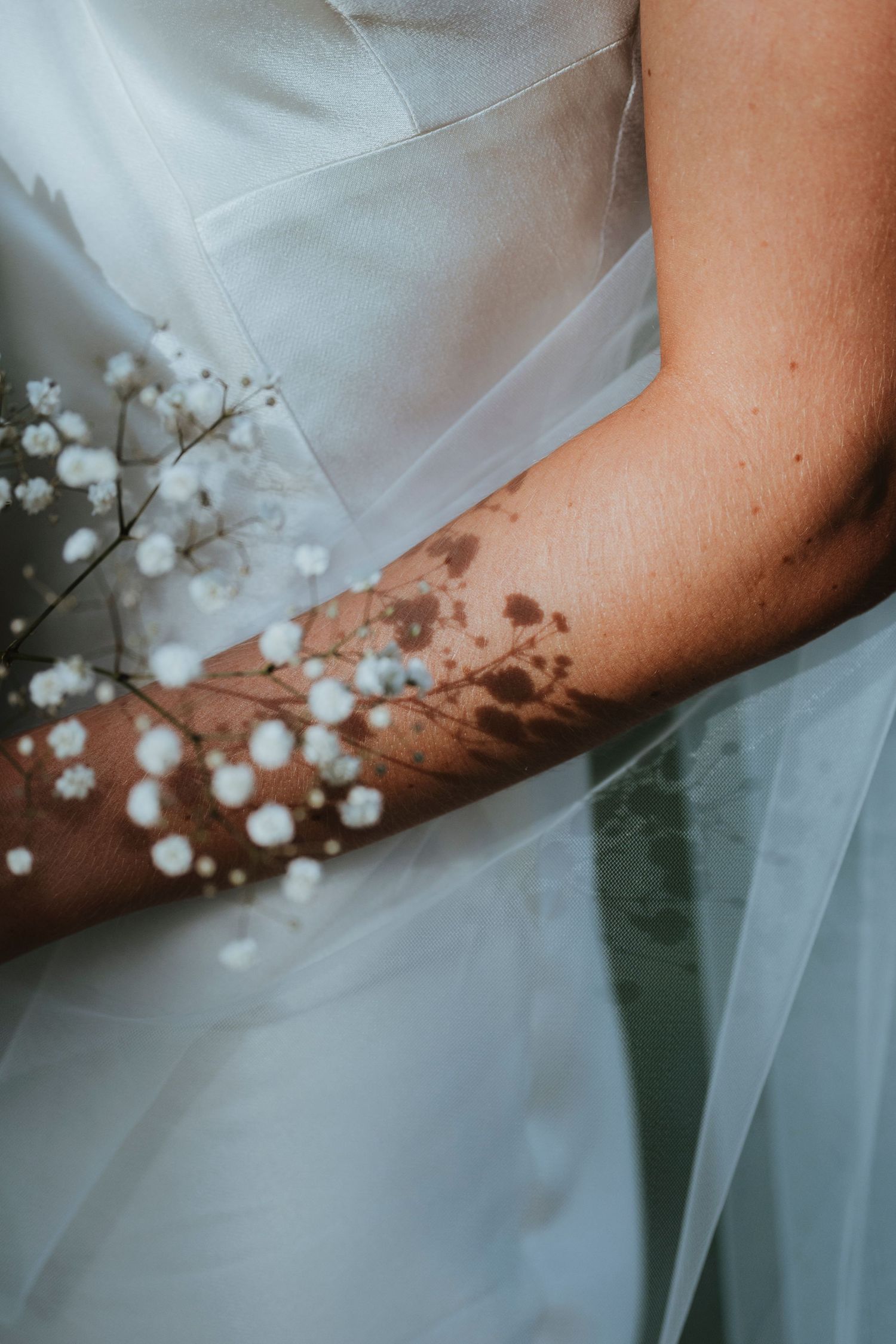 Close-up of a bride in white dress holding delicate baby's breath flowers, with soft shadows cast on her arm.
