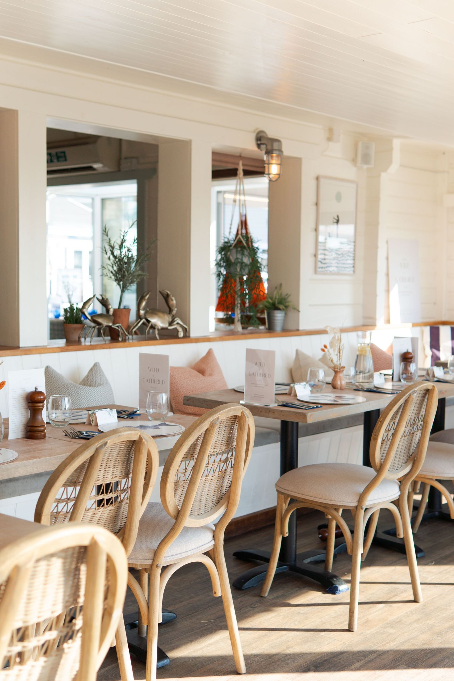 Bright restaurant interior with rattan chairs, set wooden tables, and large mirrors reflecting natural light.