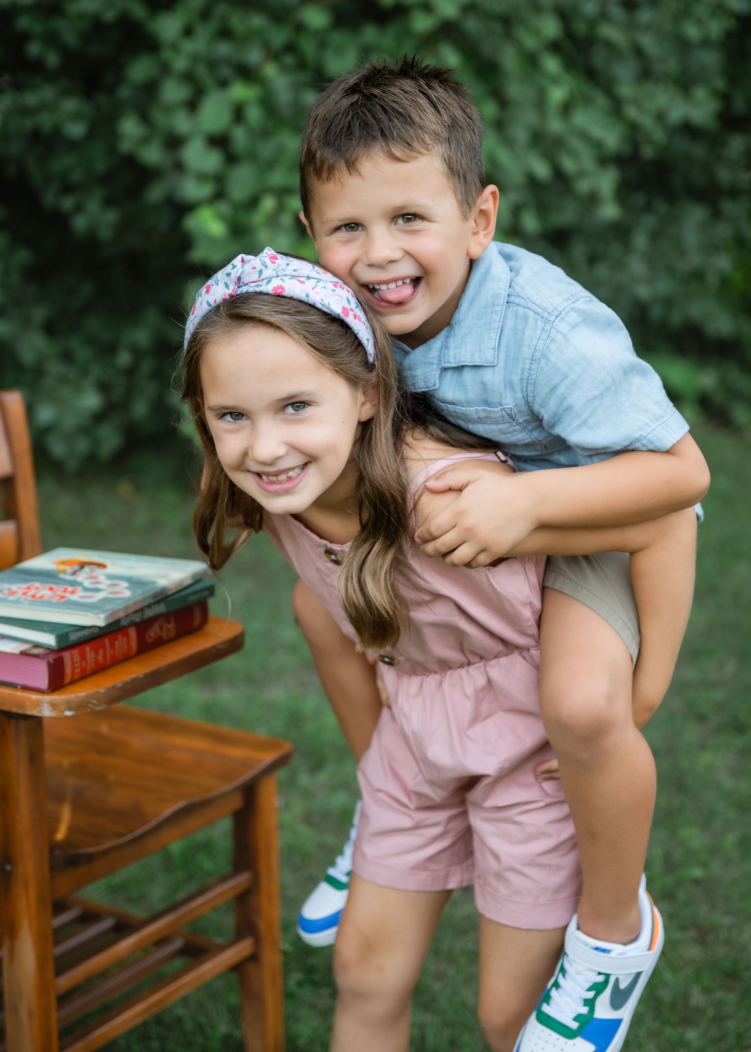 Two happy children pose outdoors, a girl in pink romper giving a boy in blue shirt a piggyback ride near a wooden chair.