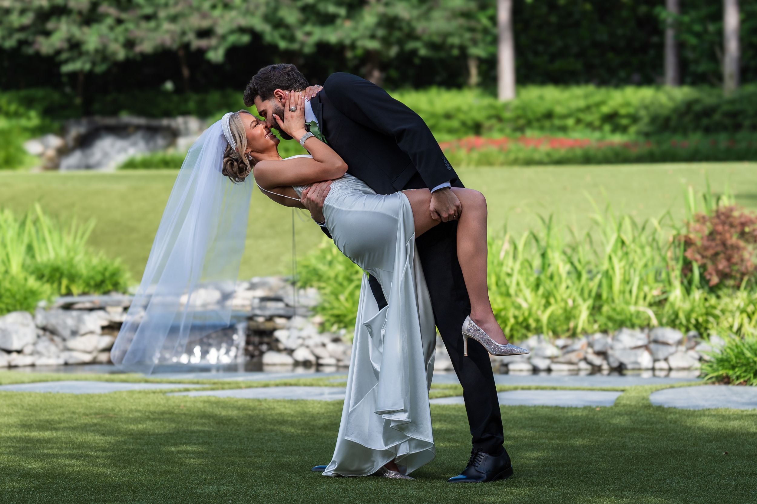 Bride and groom share a romantic dip kiss on lush green lawn during outdoor wedding ceremony.