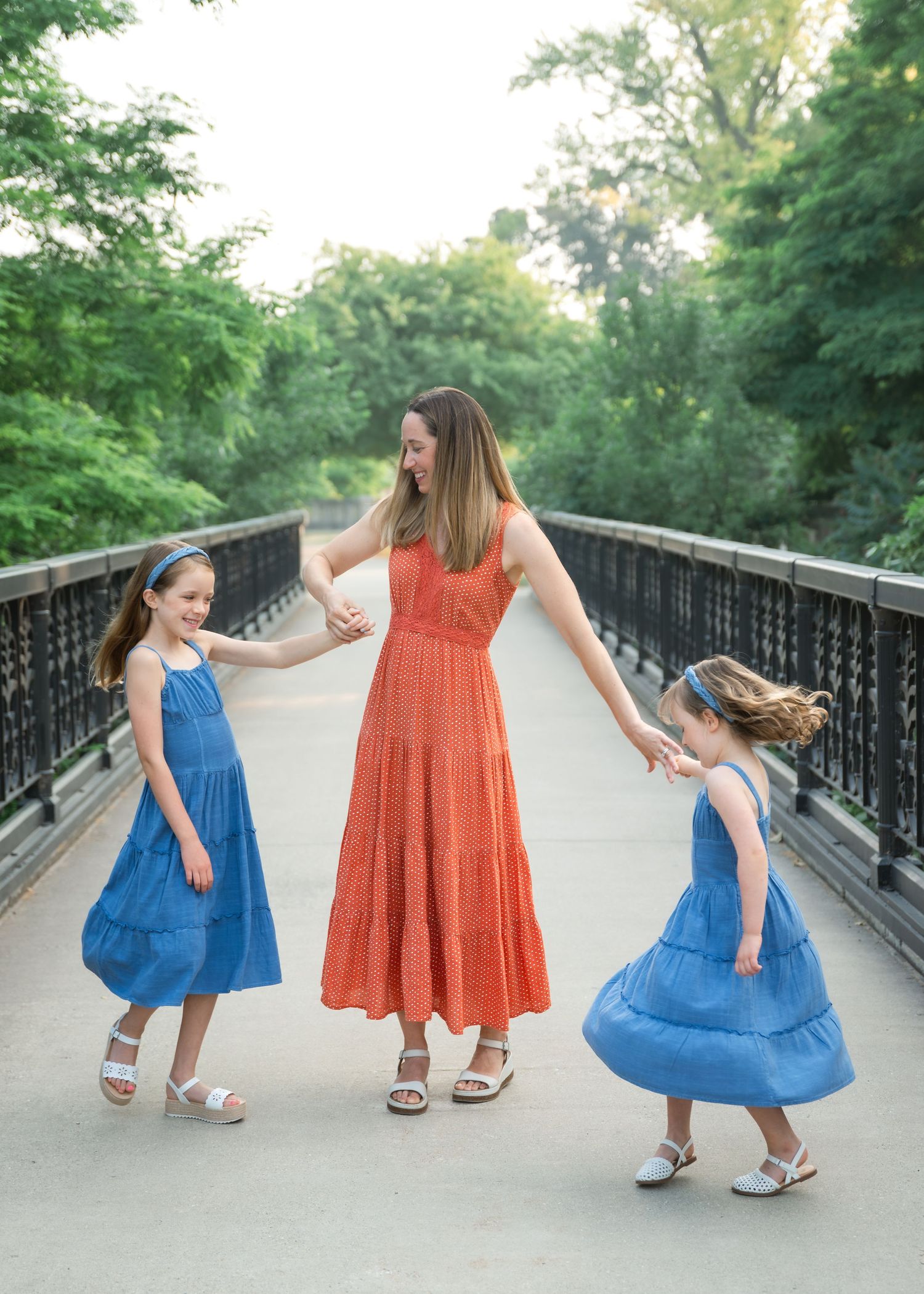 A mother in an orange dress holds hands with her two daughters in blue dresses on a bridge surrounded by green trees.