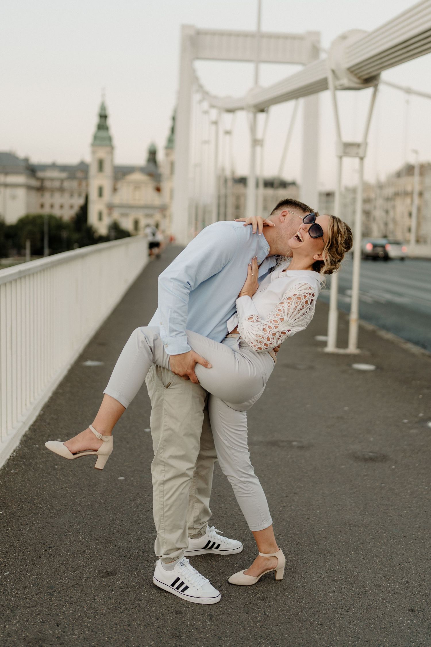 Couple sharing a romantic kiss on a bridge in Budapest, with the city skyline visible in the background.