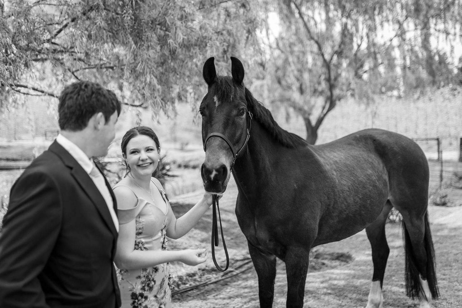 Couple smiling with a dark horse outdoors near trees in a black and white wedding photo.