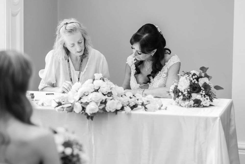 Black and white photo of two women signing wedding documents at a flower-adorned table during a ceremony.
