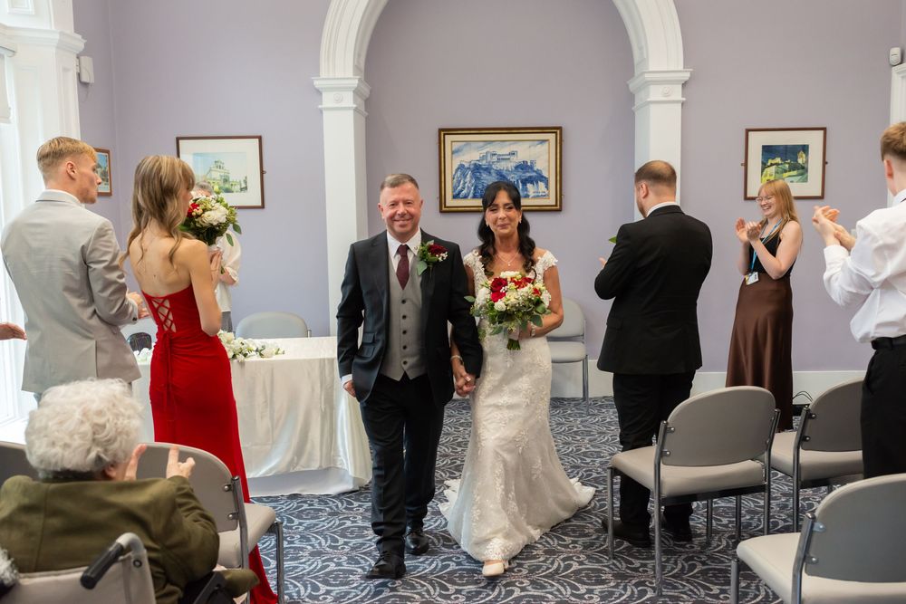 Bride and groom walk down the aisle at an elegant indoor wedding ceremony with guests applauding.