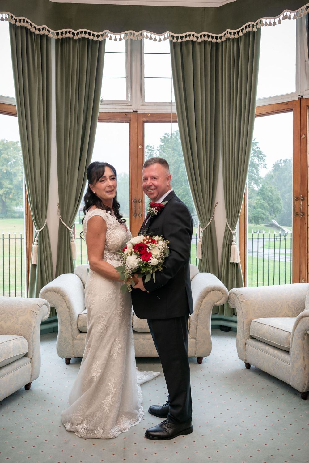 Bride and groom pose together holding a red and white bouquet in an elegant room with green curtains.