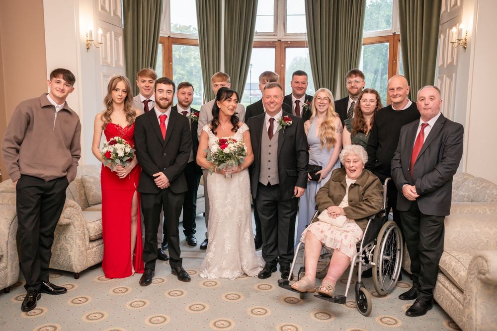 A wedding group photo with bride, groom, and family members posing indoors, including an elderly woman in a wheelchair.