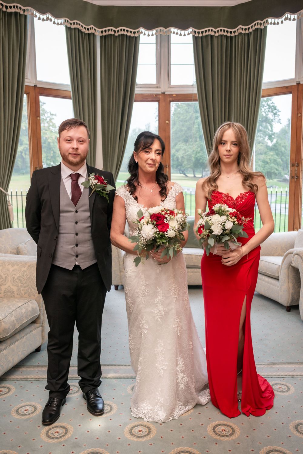 Wedding party poses indoors, bride in lace gown holds bouquet, groom in suit, bridesmaid in red dress.