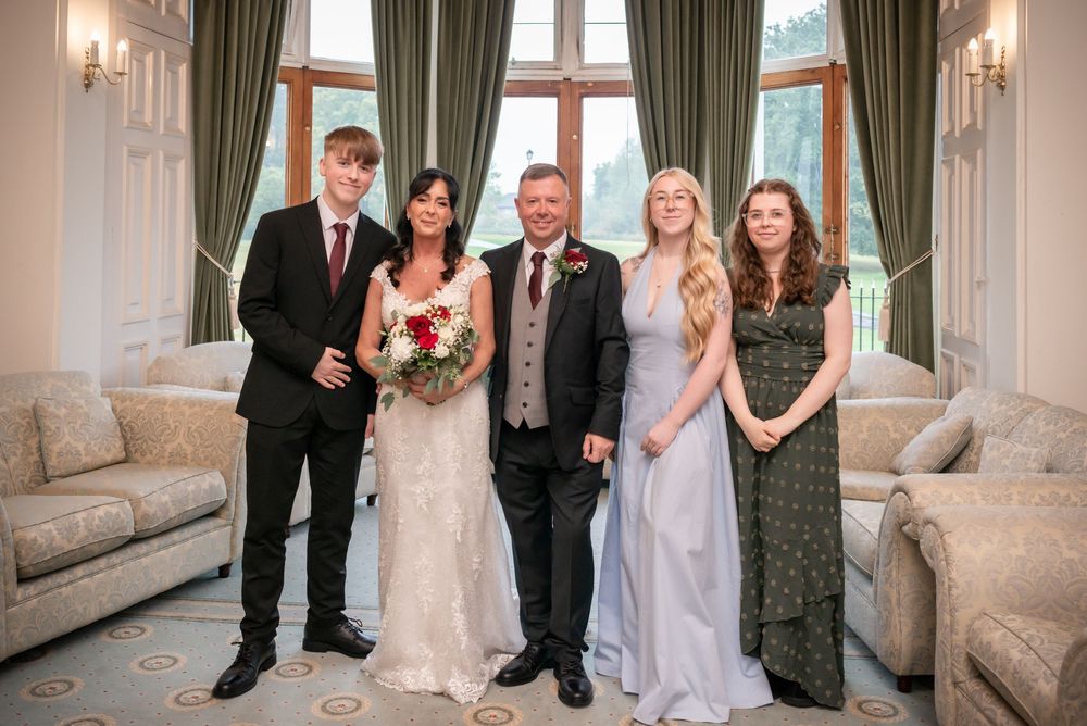 Wedding party of five posing indoors, bride in white holding red bouquet, groom in suit, flanked by three young adults.