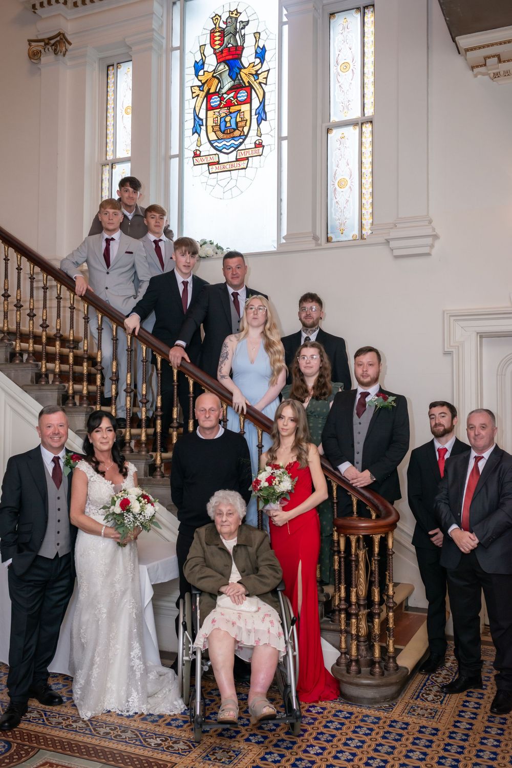 Large wedding family group photo on ornate staircase with stained glass coat of arms window in elegant venue.