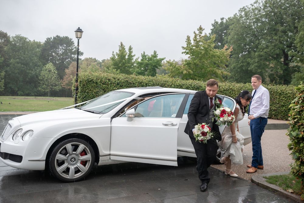 Bride and groom arriving at wedding venue in a white Bentley luxury car on a rainy day.
