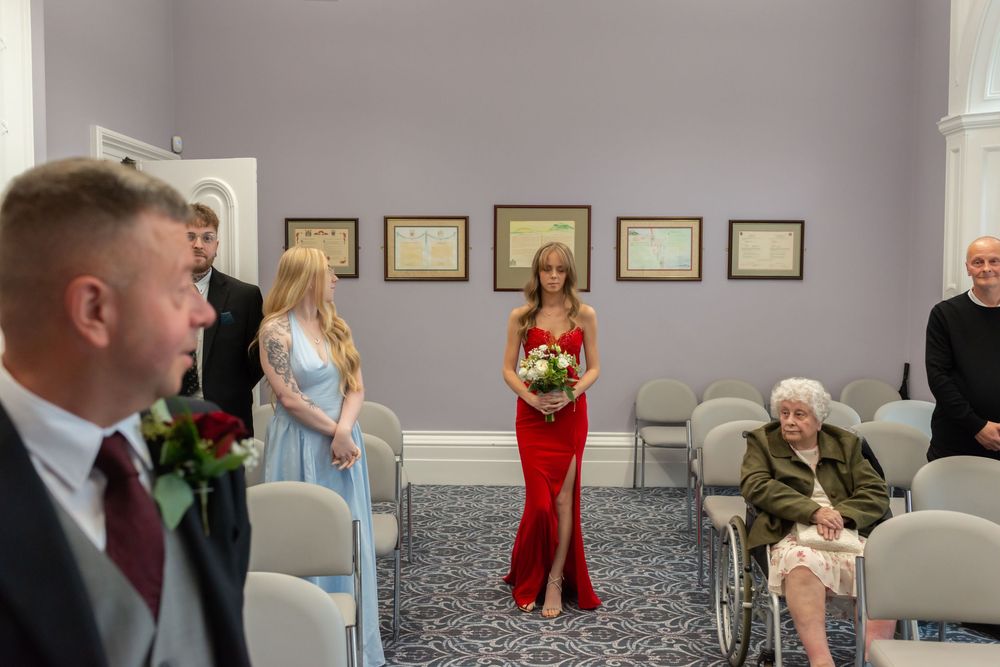Bridesmaid in red gown holds bouquet as guests gather in elegant ceremony room with elderly guest in wheelchair.