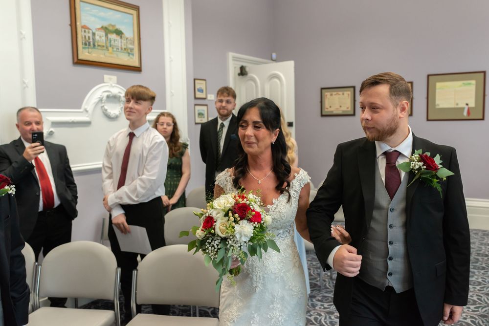 Bride in lace gown holding red and white bouquet walks down aisle with groom in gray suit at elegant wedding ceremony.
