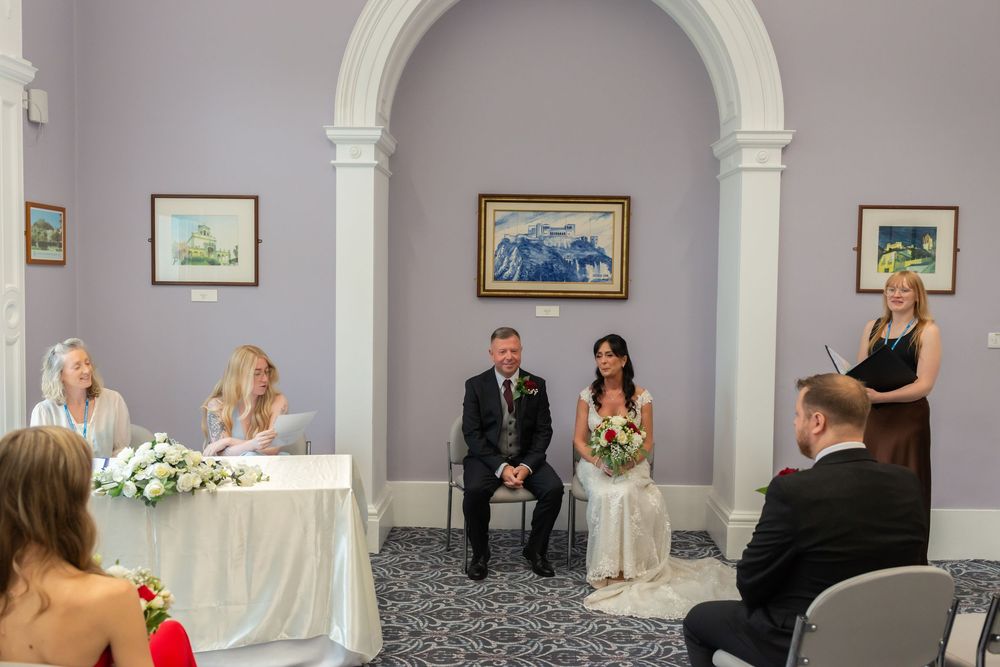 Couple seated at civil wedding ceremony in elegant purple room with officiant at podium and registrar at table.