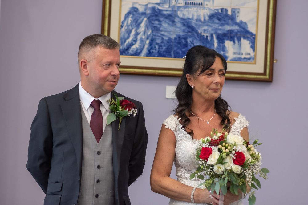 A bride holding a red and white bouquet stands beside her groom in a gray suit with a red boutonniere at their wedding ceremony.