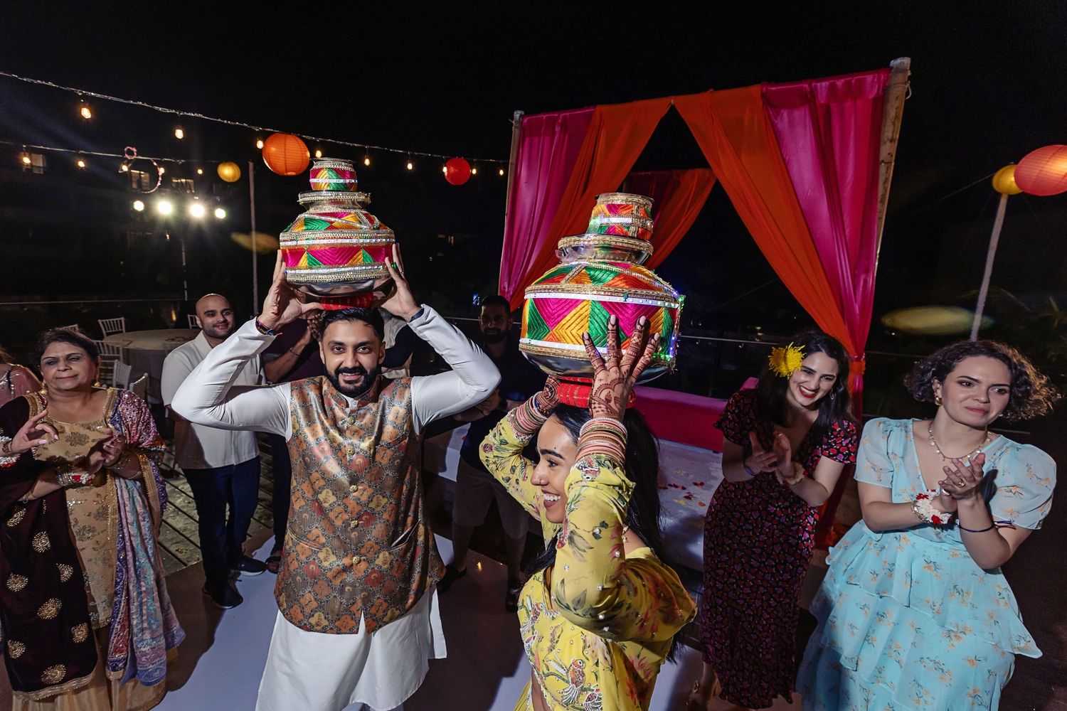 Colorful Indian wedding celebration with couple balancing decorated pots on heads, guests dancing under string lights at night.