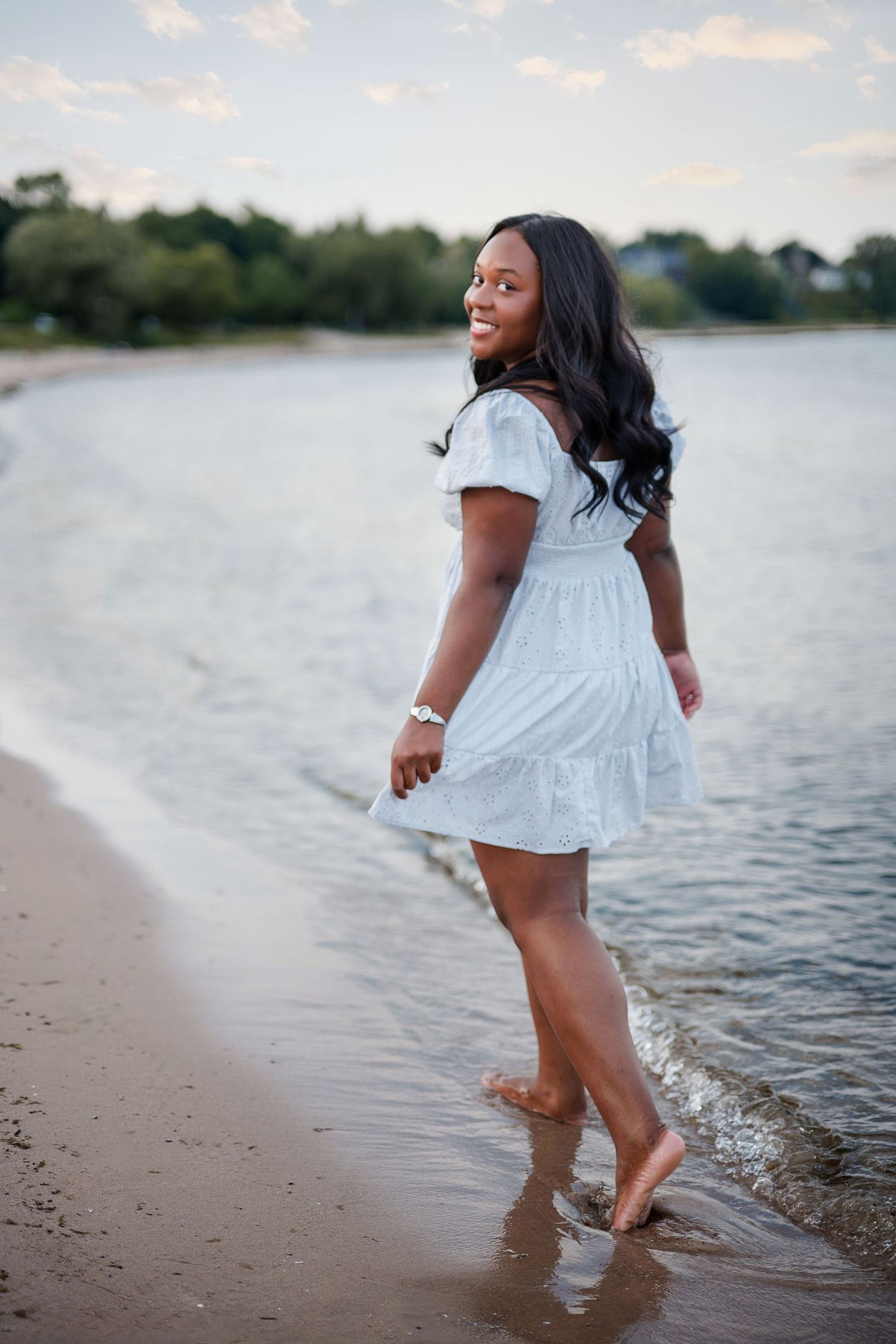 Young Black woman in white dress smiling on beach shoreline, barefoot with waves at her feet on a cloudy day.