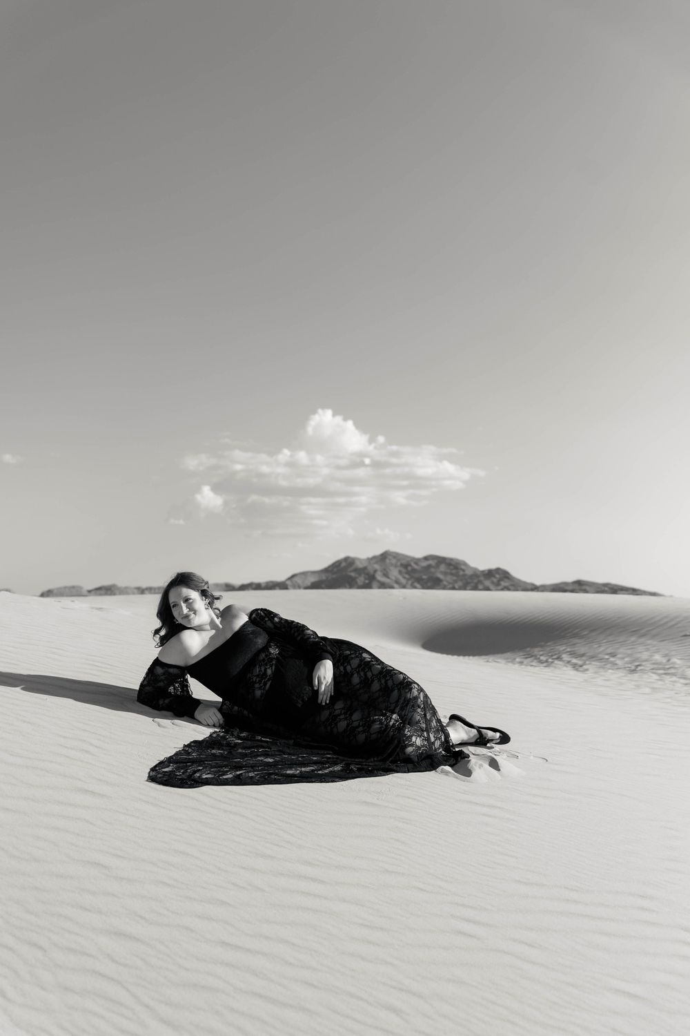 Black and white photo of woman in black lace gown posing on white sand dunes.