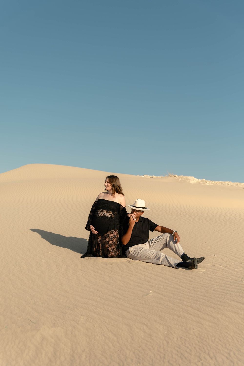 Couple sitting back-to-back on sandy desert dunes under clear blue sky, woman in black lace dress.