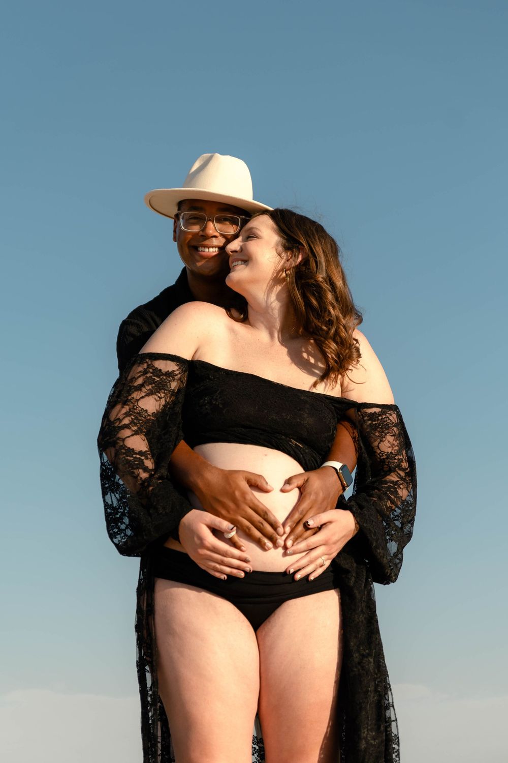 Man in white hat embracing pregnant woman from behind, both making heart shape over baby bump outdoors.