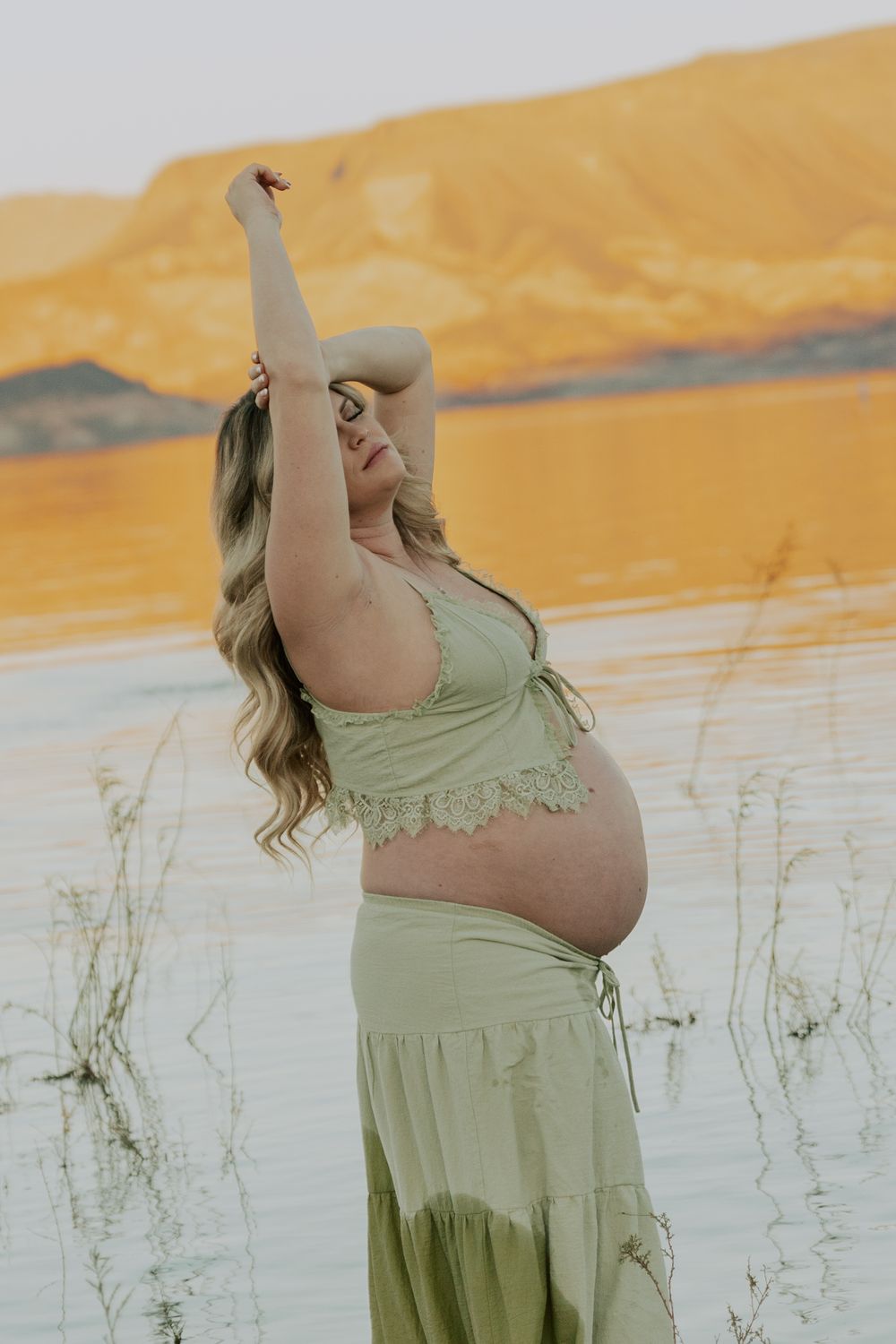 Pregnant woman in green two-piece outfit poses by golden lake at sunset.