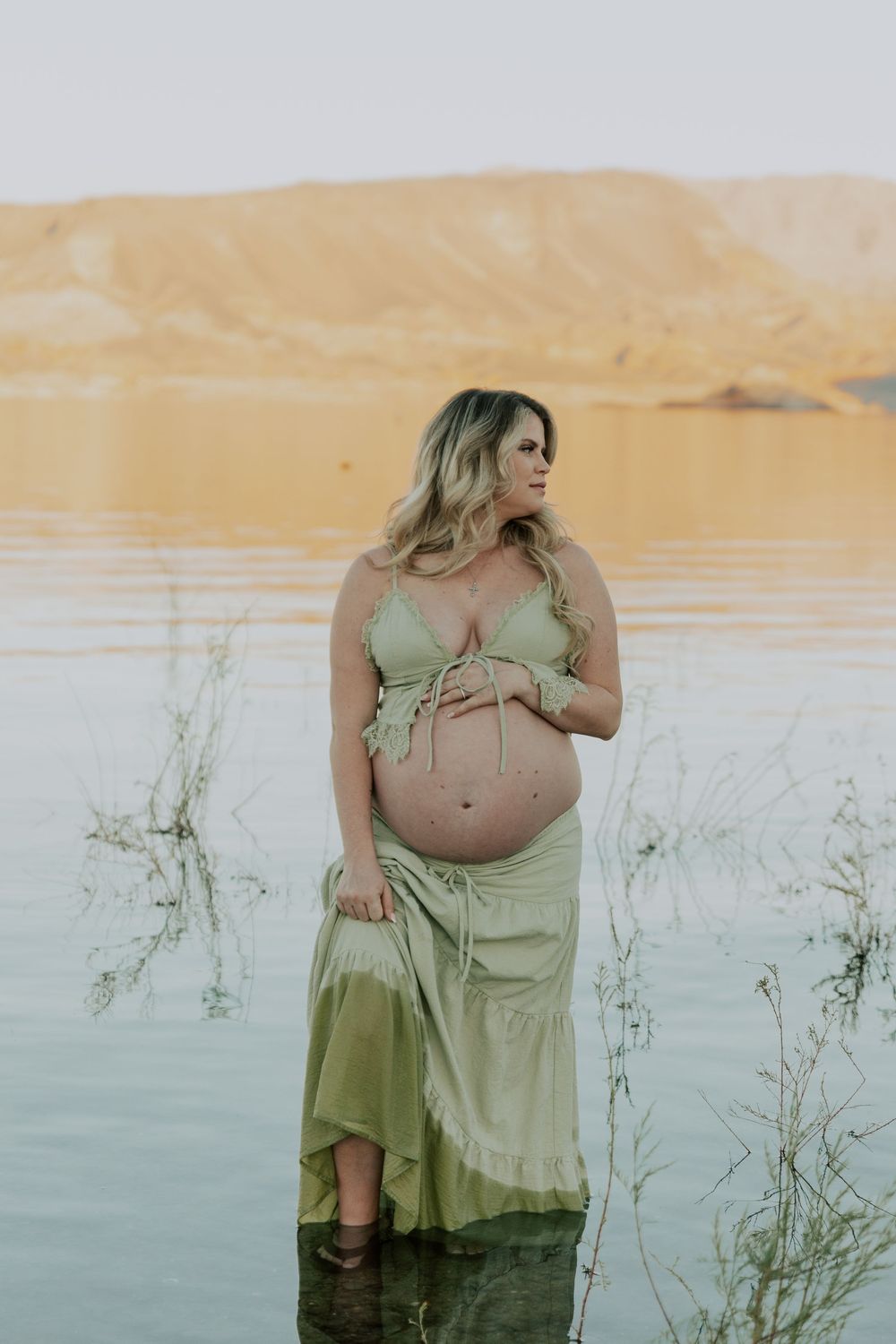 Pregnant woman in sage green dress stands in lake with golden sand dunes behind.