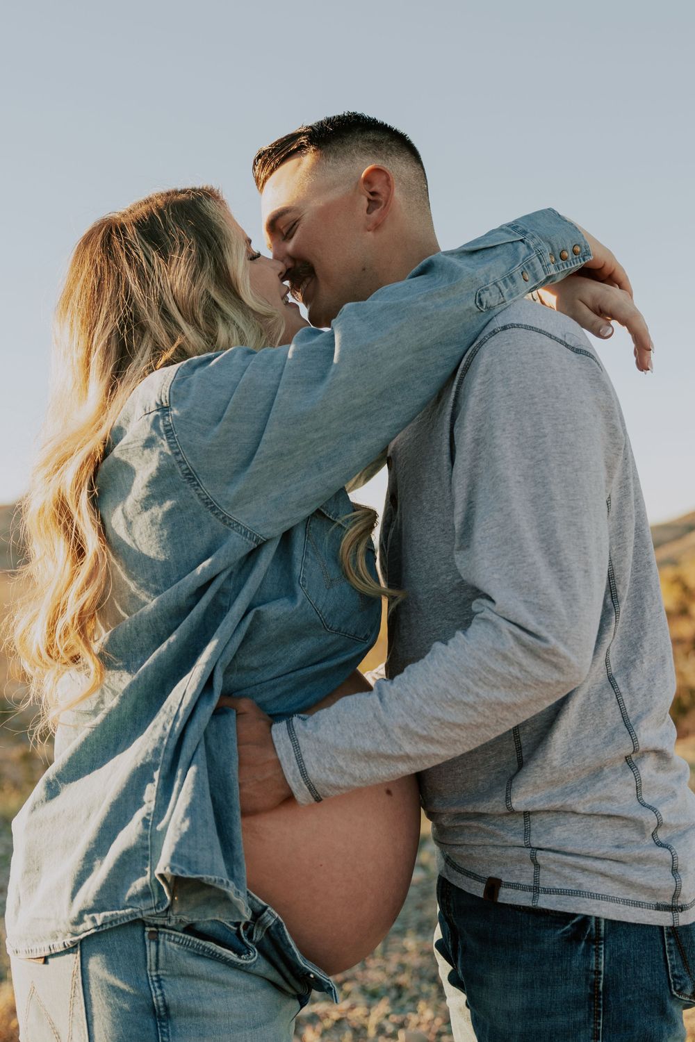 Couple shares a kiss outdoors in denim and gray outfits during maternity shoot.