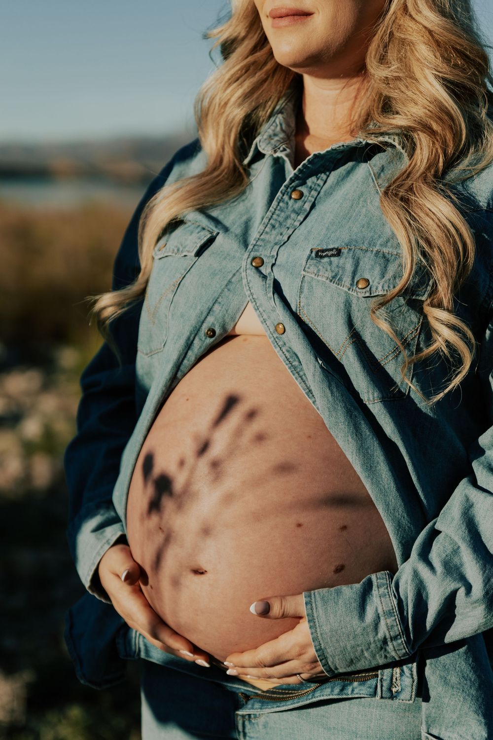 Close-up of pregnant belly with floral shadow through open denim shirt outdoors.