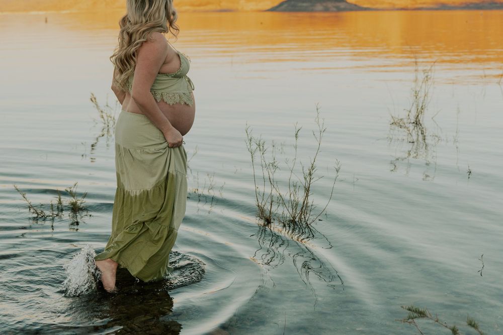 Pregnant woman in sage green dress stands in shallow water at golden sunset.