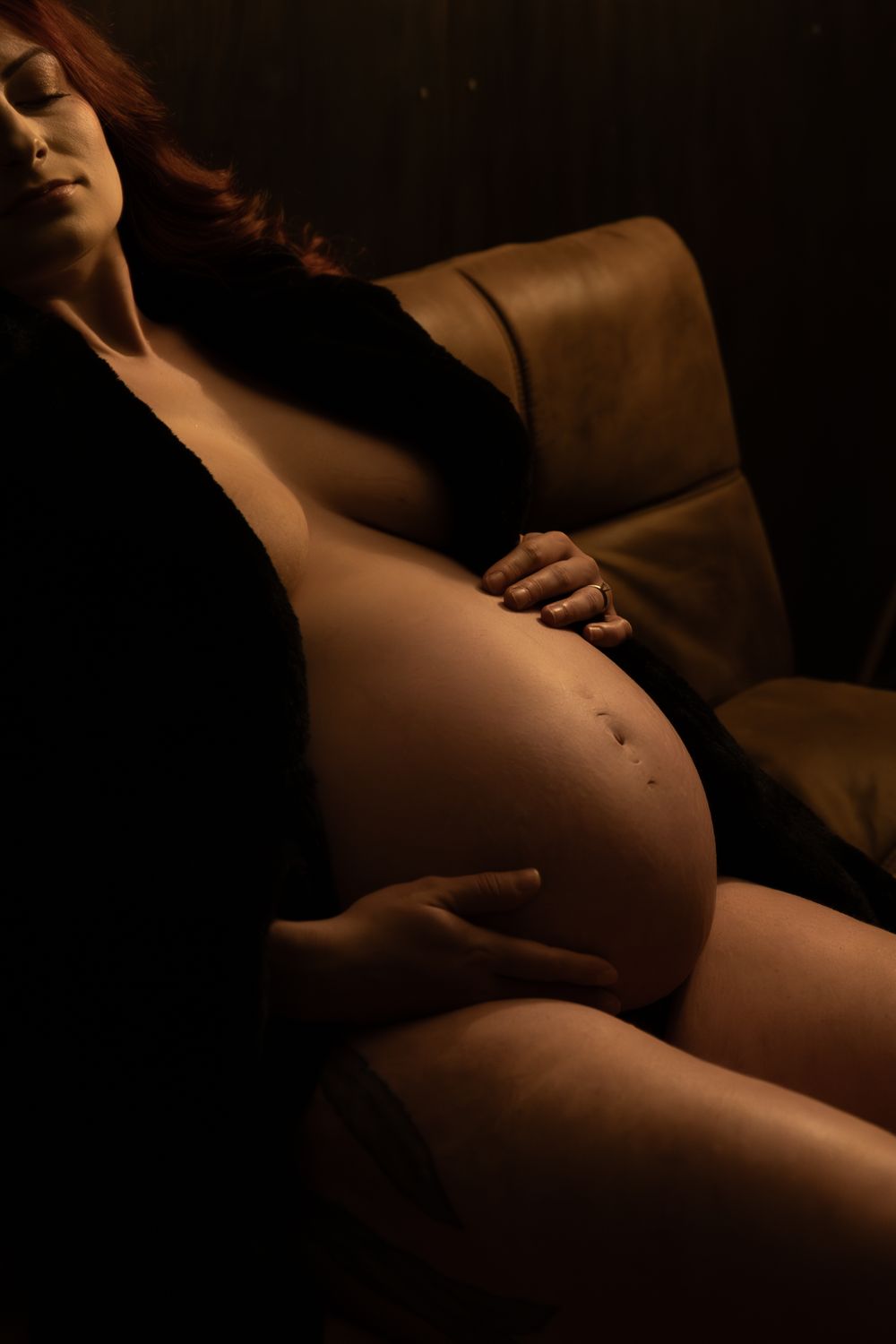 A pregnant woman with red hair cradles her bare belly while reclining in a chair in dramatic low lighting.