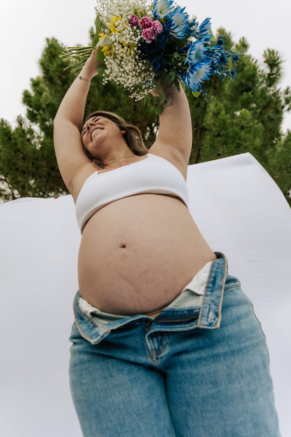 Pregnant woman in white crop top and jeans holds colorful bouquet overhead outdoors.