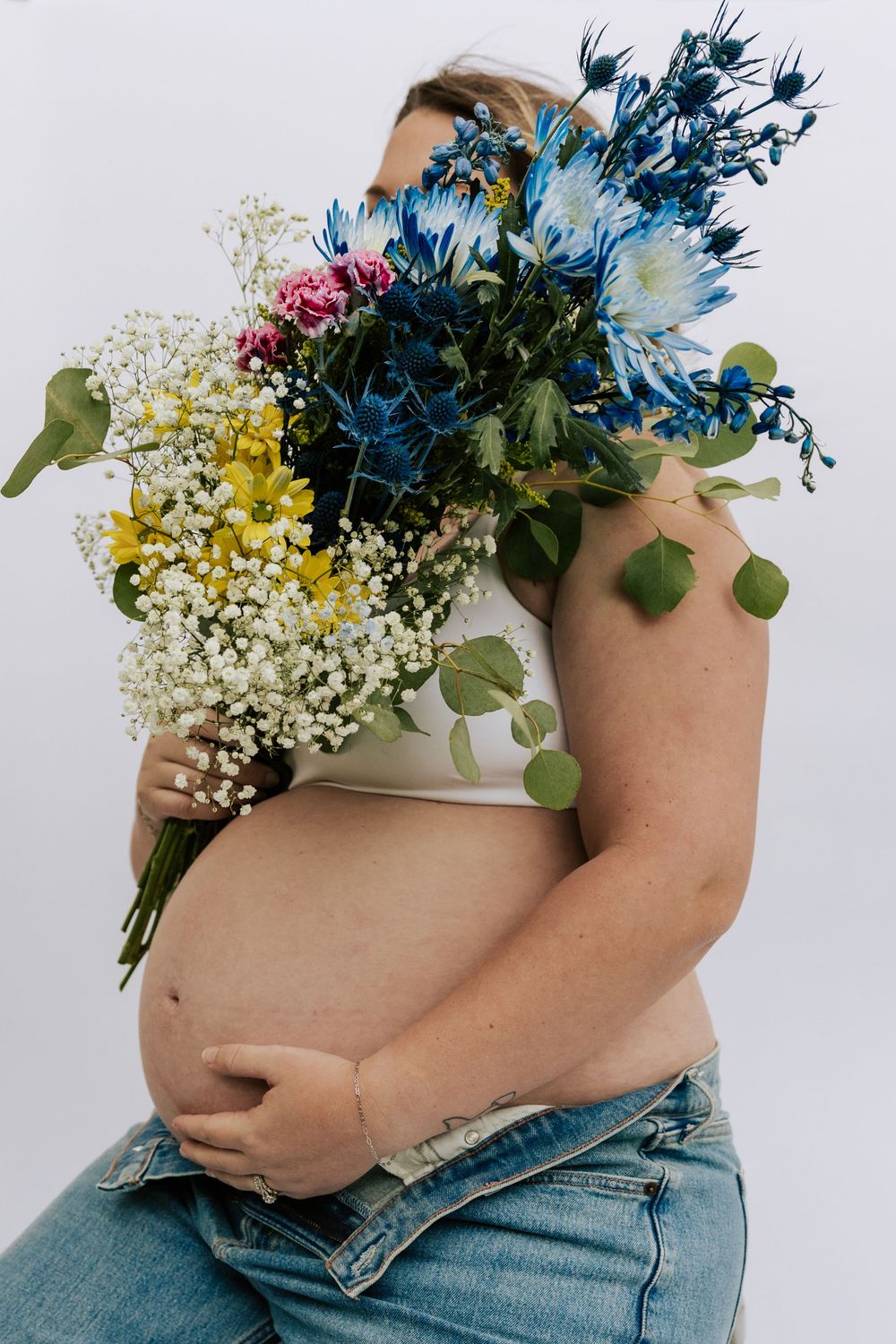 Pregnant woman in white crop top holds vibrant blue and yellow flower bouquet.