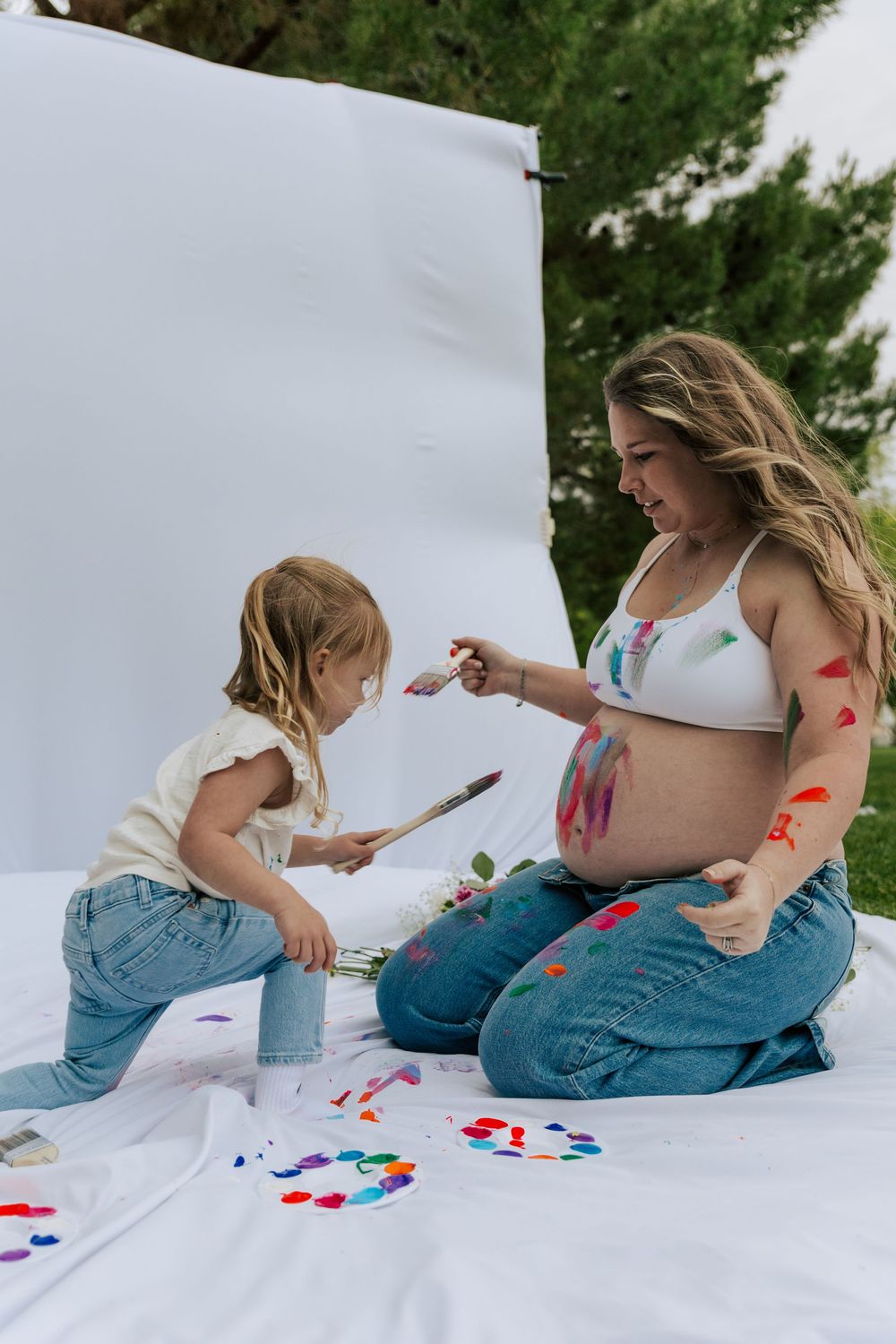 Pregnant woman and young daughter painting each other with colorful body paint on white backdrop outdoors.