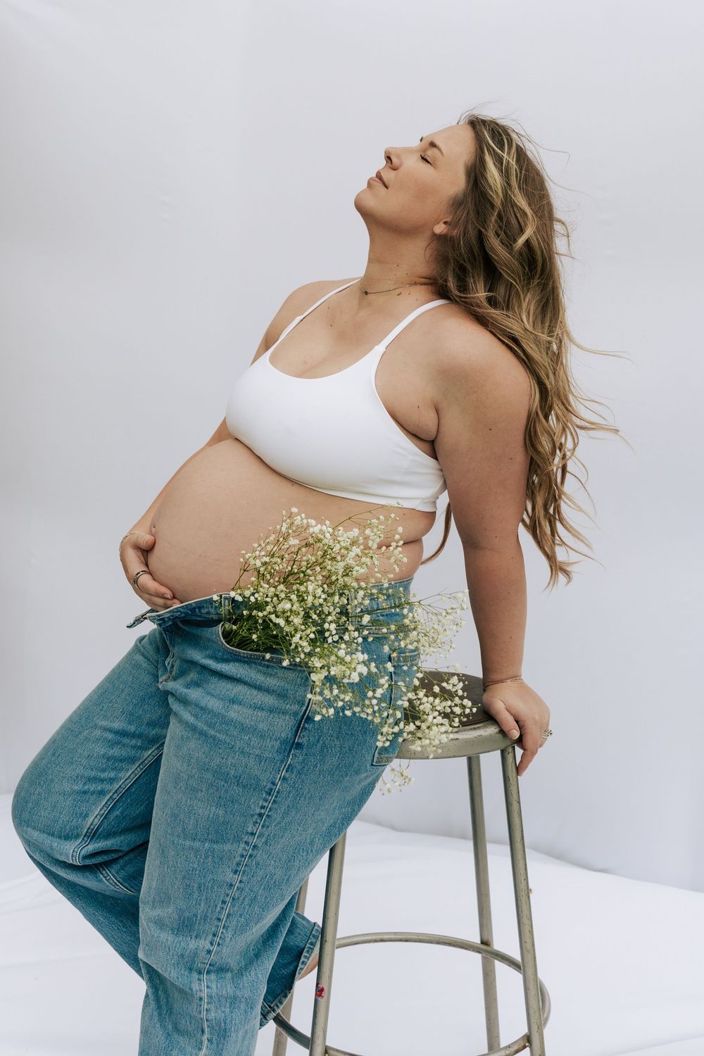 Pregnant woman in white crop top and jeans leaning back on stool with baby's breath flowers in waistband.