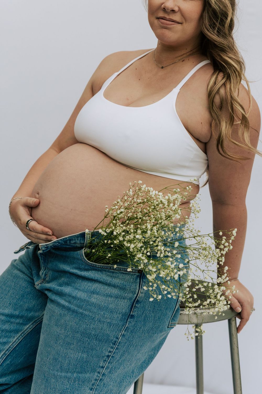 Pregnant woman in white crop top holding baby bump with baby's breath flowers tucked in open jeans waistband.