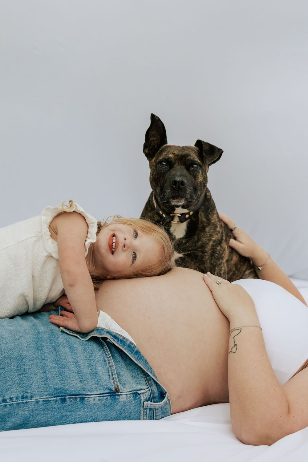 Young girl resting head on pregnant mothers bare belly while brindle dog sits behind them on white backdrop.