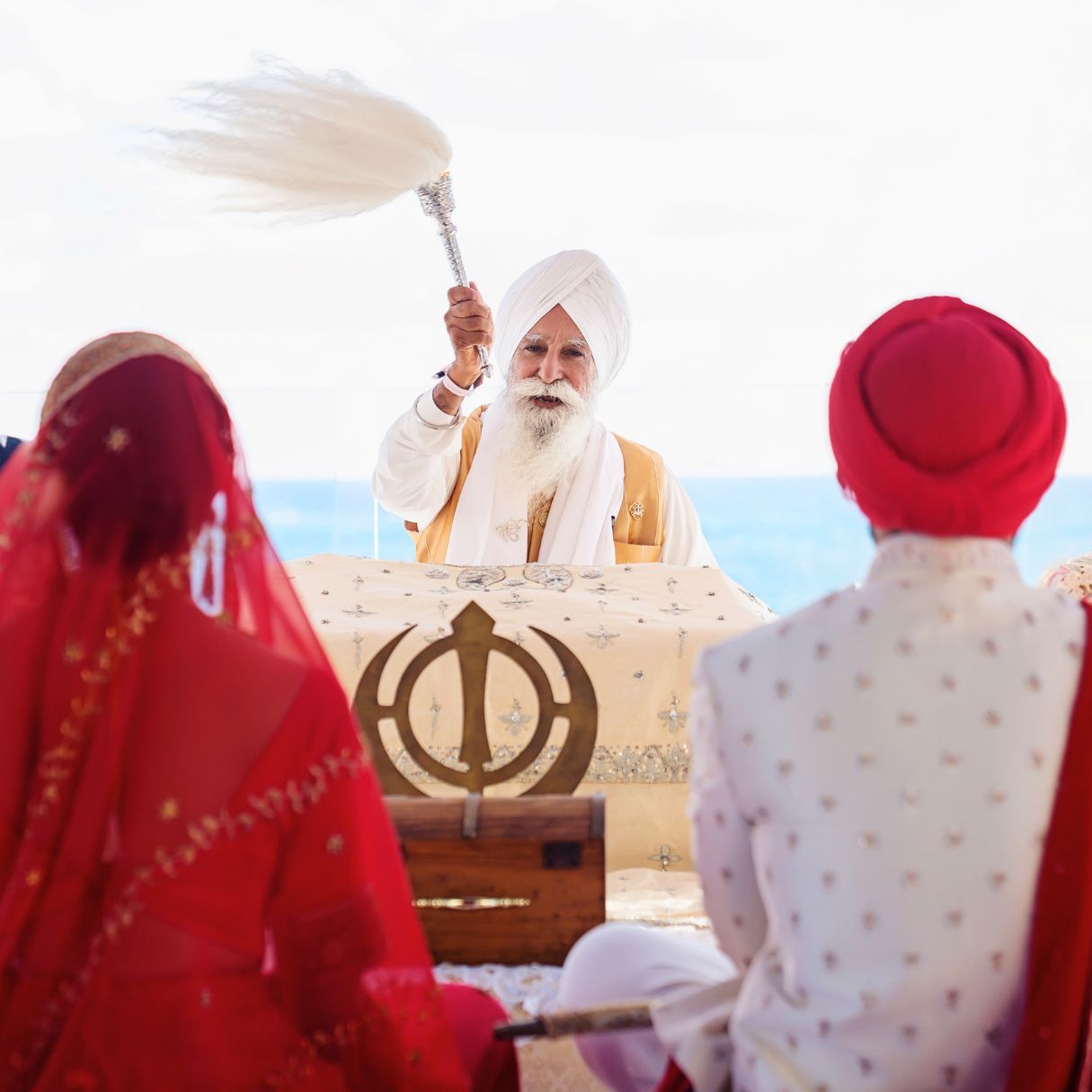Sikh wedding ceremony with priest waving chaur sahib over bride and groom before Khanda symbol outdoors.
