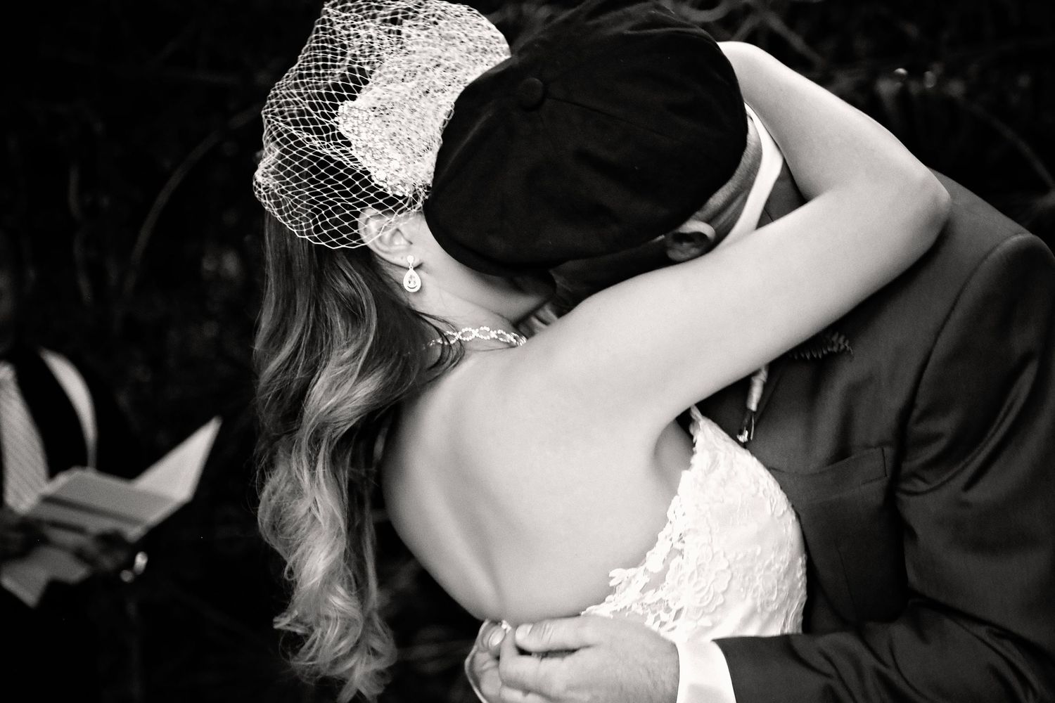 Black and white photo of a bride in a birdcage veil embracing a groom in a cap and suit during an outdoor wedding ceremony.