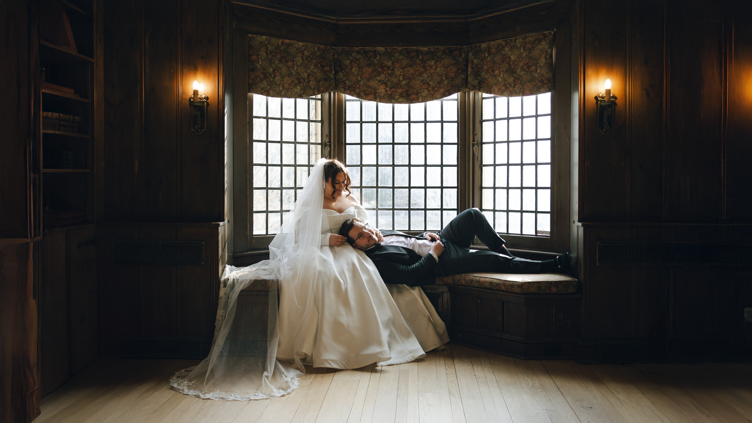 A bride in a flowing white gown sits gracefully by a large bay window in a dimly lit vintage room.