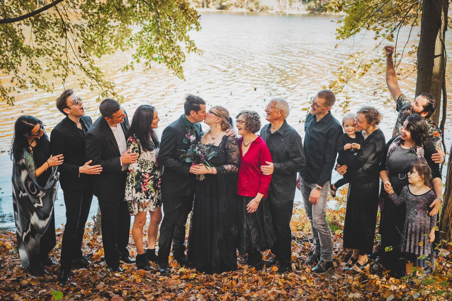 A large family group poses outdoors near a lake surrounded by autumn foliage during a fall celebration at Seneca Creek State Park