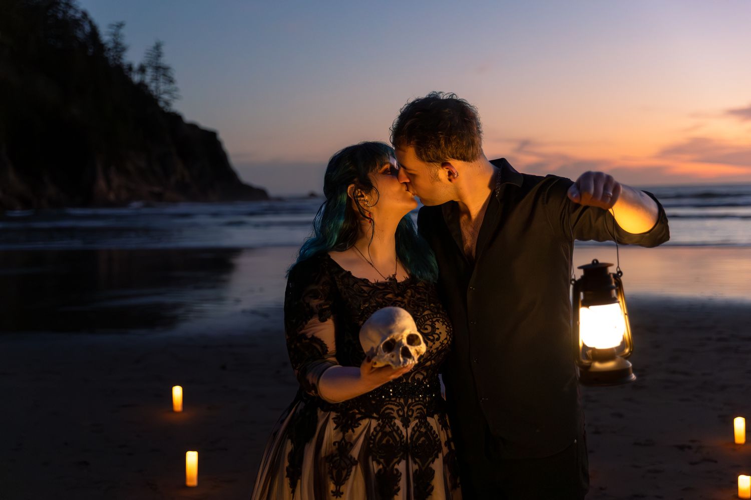 Couple in elegant dark attire sitting together on a rocky beach surrounded by lush evergreen trees at dusk.