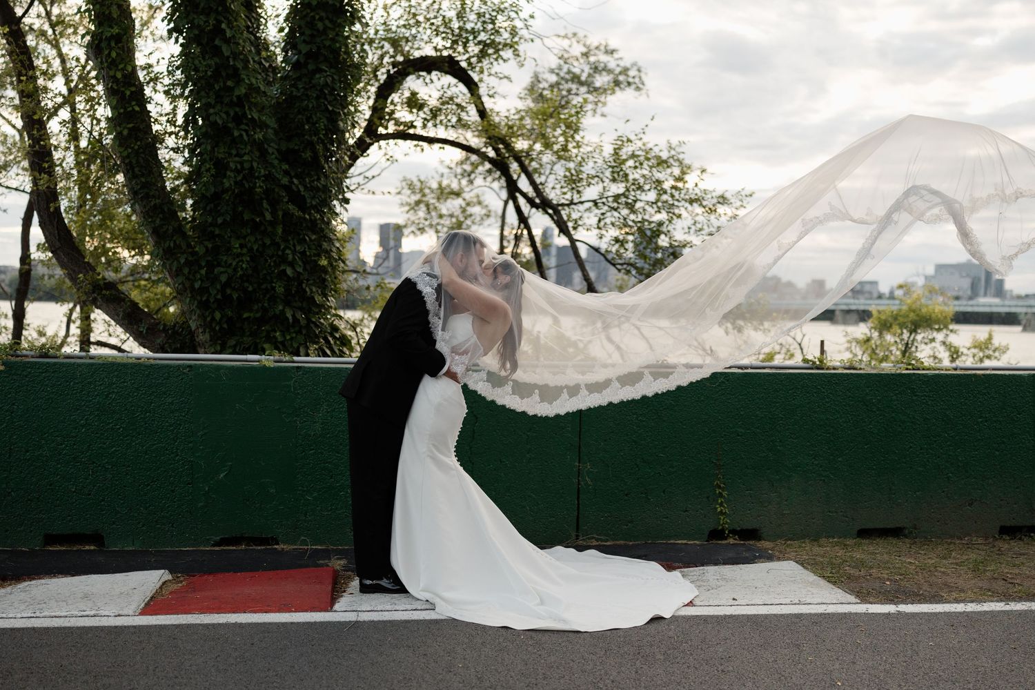 Bride and groom share a romantic dip kiss outdoors, with wedding veil flowing in the wind near a waterfront.