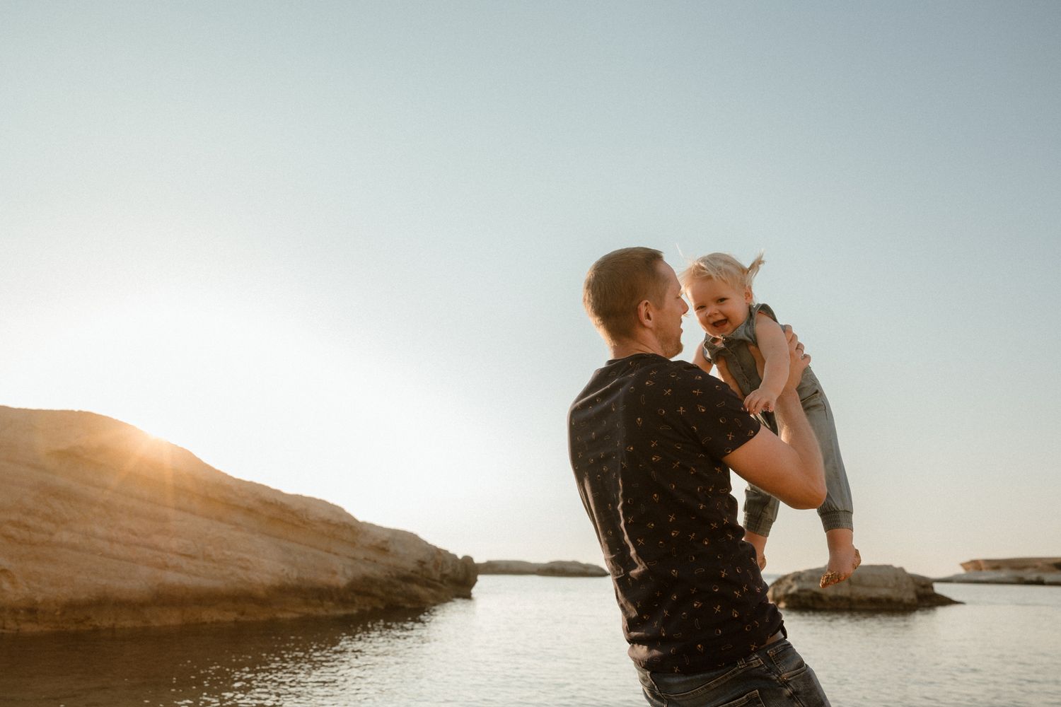 Father lifting smiling baby at sunset by rocky shoreline, golden hour family moment outdoors.