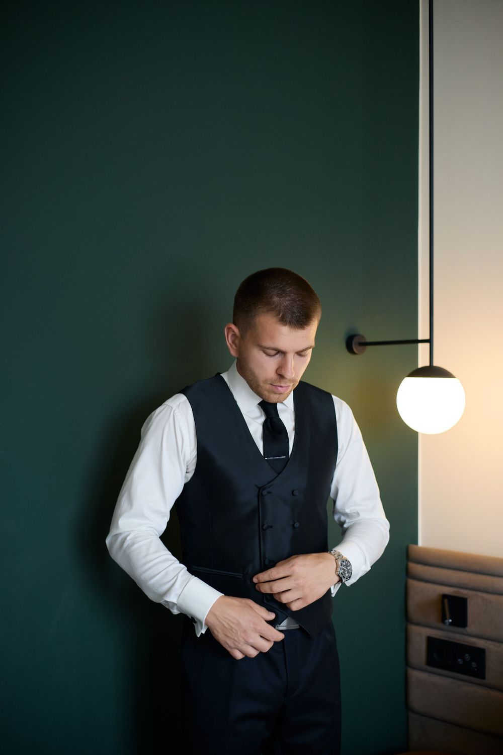 Stylish groom in a dark vest and tie buttoning his jacket against a green wall with modern lamp.
